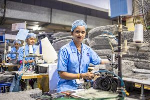 Female textile worker operates a sewing machine in a busy factory setting.
